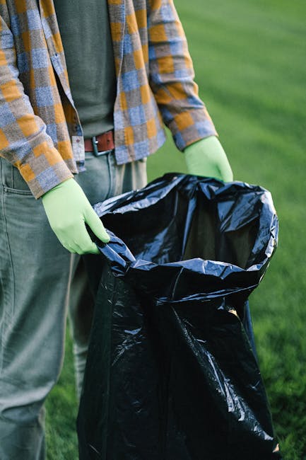 A person dressed in orange work clothing and white shoes is holding two large blue plastic trash bags filled with waste, standing on a smooth, light grey concrete floor against a plain, light-colored 