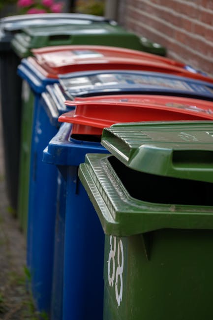A collection of overflowing rubbish bins and scattered waste materials situated outdoors on a paved area next to a parking space, with a silver car parked nearby. The central grey mixed paper and card